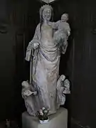 Photographie en couleurs d'une statue en pierre représentant une mère portant un enfant à son bras gauche, entourée de musiciens, dans un intérieur couvert de boiseries.