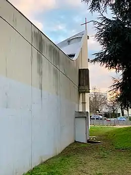 Vue de côté d'un bâtiment en béton, avec des traces d'humidité ; le bâtiment se prolonge avec une partie montante surplombée d'une croix.