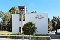 Vue de face d'un bâtiment carré en béton, surmonté d'une cloche dans un cube en béton ; sur la façade est écrit "église Saint Pierre Chanel". Au premier plan, un buisson et des poteaux pour accrocher des vélos. En arrière plan, des arbres et quelques voitures.