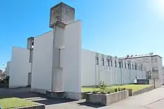 Vue de côté d'un long bâtiment en béton, avec une haute façade blanche surmontée d'un cube dans lequel se trouve une cloche. Le reste du bâtiment, cubique, comporte plusieurs fenêtres. Du gazon et une route entourent le bâtiment.