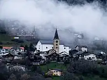 Vue de l'église Saint-Romain et de son voisinage. Une couche de nuage embrume le haut du village, à l'arrière plan.