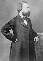 Photographie en noir et blanc et de profil d’un homme barbu, portant une longue veste et un nœud papillon, se tenant debout, le poing droit sur sa hanche et le bras gauche sur un dossier de chaise