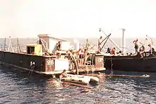 Photographie couleur argentique. En mer un pneumatique au premier plan. Deux barges à couple chargent un morceau de bois encore dans l'eau.