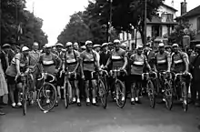Photographie en noir et blanc d'une équipe au départ d'une course cycliste, chaque coureur se tenant debout à côté de son vélo.