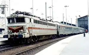 La photographie couleur montre une locomotive CC 40100, couleur inox et rouge, tractant des voitures PBA, fourgon-générateur en tête, à quai en gare de Paris Nord.