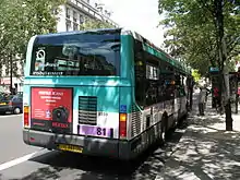 Vue de trois quarts de l'arrière d'un bus blanc et vert, avec un numéro 81 sur fonds violet, arrêté à l'ombre dans une rue bordée d'arbres.