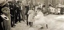 Photographie noir et blanc. Cade champêtre d'un homme genou à terre devant une fillette recevant une couronne en présence d'une assemblée.