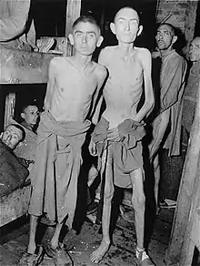 Au premier plan d'une photo en noir et blanc, trois hommes émaciés posent debout devant des lits superposés dans lesquels sont couchés d'autres prisonniers.