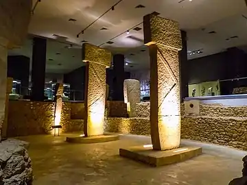 Deux pierres levées, chacune surmontée d'une autre pierre donnant une forme de T. Elle sont entourées de petits murs de pierres. Le tout est sous un plafond plat et illuminé par des projecteurs électriques.
