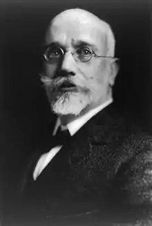 Photographie en noir et blanc d'un homme chauve portant de petites lunettes, une barbe et une large moustache.