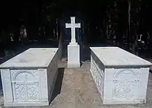 Photographie en couleurs de deux tombeaux blancs situés de part et d'autre d'une croix.