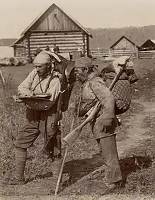 Photographie en noir et blanc de deux hommes avec des fusils devant des habitations.