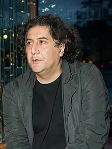 Homme aux cheveux bruns, mi-longs et frisés, au nez légèrement bombé, portant un t-shirt noir et une veste gris foncée.