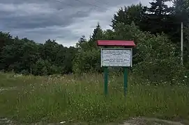 Un écriteau en bois à hauteur d'homme planté dans une prairie entourée de forêts de conifères. L'écriteau porte trois inscriptions noires sur fond blanc, en ukrainien, hébreu et anglais ; celle en anglais indique : Mass grave of Boryslaw Jews murdered by the "Nazis" at 28.11.1941 God rest their soul!