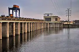 Vue du barrage de Kakhovka en 2013 : les arches du déversoir traversent le cours d'eau. À l'autre bout du barrage, le bâtiment des turbines se trouve sur l'ouvrage. Des lignes haute tension enjambent le fleuve. Une grue bleue est positionnée sur le barrage.