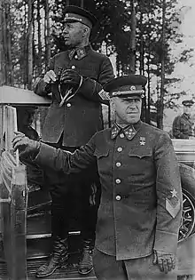 Photo noir et blanc avec deux hommes en uniforme au premier plan, l'un sur le marche-pied d'une voiture et tenant des jumelles, l'autre en dessous tenant la portière.