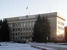 Photographie en couleurs. Bâtiment en pavé droit haut de trois et surmonté d’un drapeau ukrainien. Des conifères masquent partiellement la façade et de la neige recouvre le sol. Ciel dégagé.