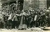 Photographie en noir et blanc d'une chanteuse en costume traditionnel accompagnée d'un orchestre militaire et chantant devant des ruines.