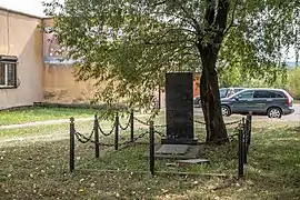 Au pied d'un arbre, une pierre tombale noire avec des inscriptions, entourée de potelets reliés par des chaînes. En arrière-plan, un bâtiment et des voitures stationnés.