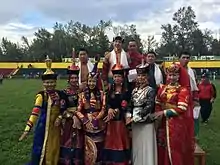 Photographie de groupe dans un stade : devant, des jeunes femmes en vêtements colorés, derrière, les lutteurs en tenue de combat.