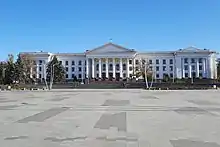 Photographie en couleurs. Au premier plan, le revêtement teinté de gris de la place vierge de toute occupation. Sur le second, la façade d’un grand bâtiment blanc de style néo-classique haut de quatre étages occupe un large espace. Quelques arbres sont visibles. Ciel dégagé en surplomb.