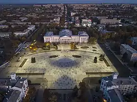 Photographie en couleurs. Vue en plongée sur la place. La place est partiellement végétalisée et peuplée de passants. Au centre de la photographie, un bâtiment blanc à quatre étages de style néo-classique barre la place sur la quasi-totalité de sa largeur. Présence d’immeubles à perte de vue autour de la place. Un ciel nocturne dégagé surplombe faiblement l’ensemble.