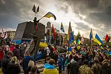 Photographie en couleurs. Une statue de Lénine est en berne, laissant son piédestal vide, au milieu d’une foule de manifestants arborant des drapeaux ukrainien et de « Secteur droit ». Un ciel chargée de nuage et de lumière recouvre la scène.