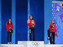 Trois biathlètes sur le podium, chacune tenant un bouquet de fleur dans les mains.
