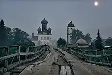 Dans une atmosphère grise et brouillardeuse, un pont de guingois ouvre la voie vers une ancienne église.