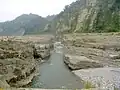 Photo couleur de falaises stratifiées beiges d'un canyon traversé par un cours d'eau (au centre). Des montagnes en arrière-plan, sous un ciel laiteux.