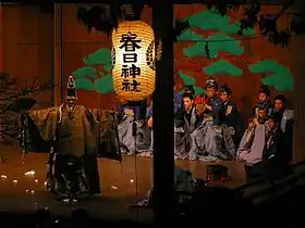 Photo couleur d'une scène de théâtre nō éclairée par une lanterne en papier. Un personnage en kimono marron se tient debout sur la scène, de dos au premier plan, et des musiciens sont assis sur deux rangées au second plan. Sur le fond marron est dessiné le feuillage vert d'un arbre.