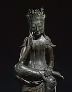 Photographie d'une sculpture d'un bouddha en métal. Il est assis, sa jambe gauche est ramenée sur le genou de sa jambe droite. Il est disposé dans un fond neutre.