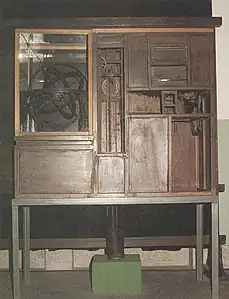 Photographie d'un meuble en bois dans lequel sont visibles des mécanismes et le cadran d'une horloge.
