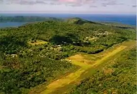 Photo aérienne montrant une large bande d'herbe verte claire, contrastant avec des arbres plus foncés, quelques nuages, des habitations clairsemées ; sur la gauche, un cratère est visible, et au fond, l'océan Oacifique.