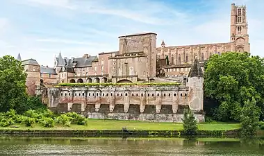 La photo couleur montre une rivière au premier plan surmontée d'un jardin fortifié et d'un château de brique rouge. En arrière-plan, un édifice religieux, lui aussi en brique, domine.