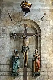 Photo couleur d'un Christ en croix encadré de femmes en prière. La croix est fixée dans une alcôve du mur à linteau roman.