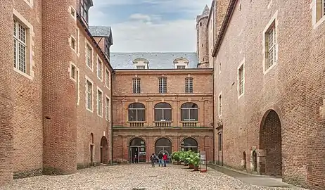 Photo couleur d'une aile de château sur cour en brique rouges. Le rez-de-chaussée comporte trois arcades vitrées de types classique à pilastres supportant une corniche en pierre. Au premier étage, le décor se répète avec une balustrade de pierre. Au second étage, trois fenêtres rectangulaires de même style ont un garde-corps en fer forgé.