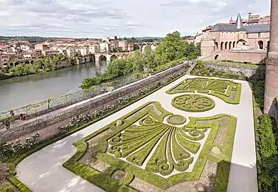 Photo couleur d'un jardin à la française de buis taillé et fleurs. En arrière plan, des escaliers et balustrades cernent deux terrasses enserrées entre une tour découronnée et des bâtiments en brique rouge de style classique.