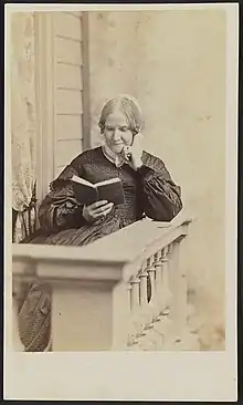  Photographie sépia d'une femme blanche d'âge mûr portant un bonnet blanc, assise et lisant sur un balcon