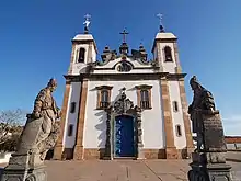 Photographie en couleurs. Vue en légère contre-plongée de l'église vue de face, avec deux statues d'apôtres sur l'entourant au premier plan.