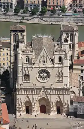Vue en couleurs d’une cathédrale. Les toitures sont visibles derrière la façade. En arrière-plan, un cours d’eau.