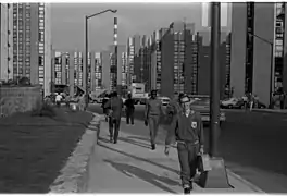 Photo en noir et blanc. Un athlète d'Allemagne de l'Est portant une mallette marche sur le trottoir ; le long de la route, des deux côtés, de nombreux immeubles hauts.