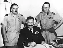 Photographie noir et blanc montrant Lukis assis à un bureau, Harry Cobby debout à sa gauche et une autre personne debout à sa droite.