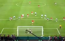Photographie en couleurs. La photographie, prise depuis les tribunes en hauteur, représente la surface de but argentine dans laquelle l'Anglais tire le penalty face au gardien argentin qui a commencé son mouvement vers la droite alors que le ballon part sur sa gauche.