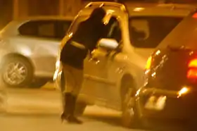Image en couleur d'une femme appuyée contre une voiture en train de discuter avec le conducteur.