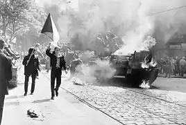 Un char d'assaut en feu au milieu de la chaussée entouré par plusieurs manifestants, dont l'un brandit un drapeau tchécoslovaque.