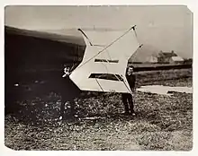 Photographie en noir et blanc de deux personnages portant un grand cerf-volant sur un terrain herbeux, légèrement accidenté.