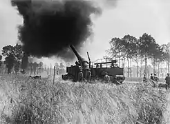 Photo noir et blanc d'un canon dans un champ arboré, avec soldats et gros panache de fumée noire.