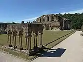 Photographie de quelques arcades romanes formant l'angle d'un cloître, tout le reste de ce dernier ayant disparu ; en arrière-plan, des ruines plus importantes.