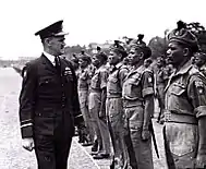 Un homme en uniforme militaire sombre avec une casquette, inspectant des troupes lors d'un défilé.